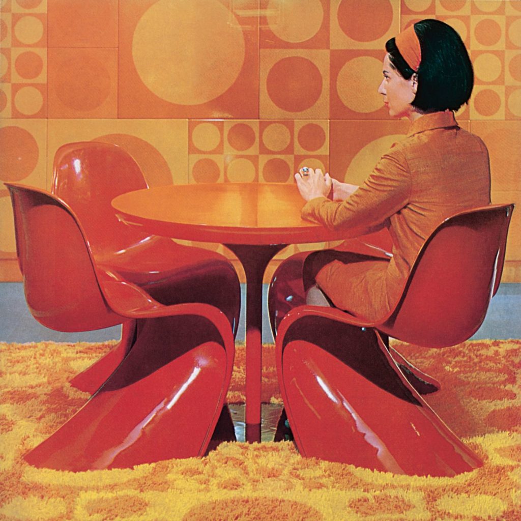 Image of a woman in late 60's fashion sitting at a table surrounded by red S-shaped chairs